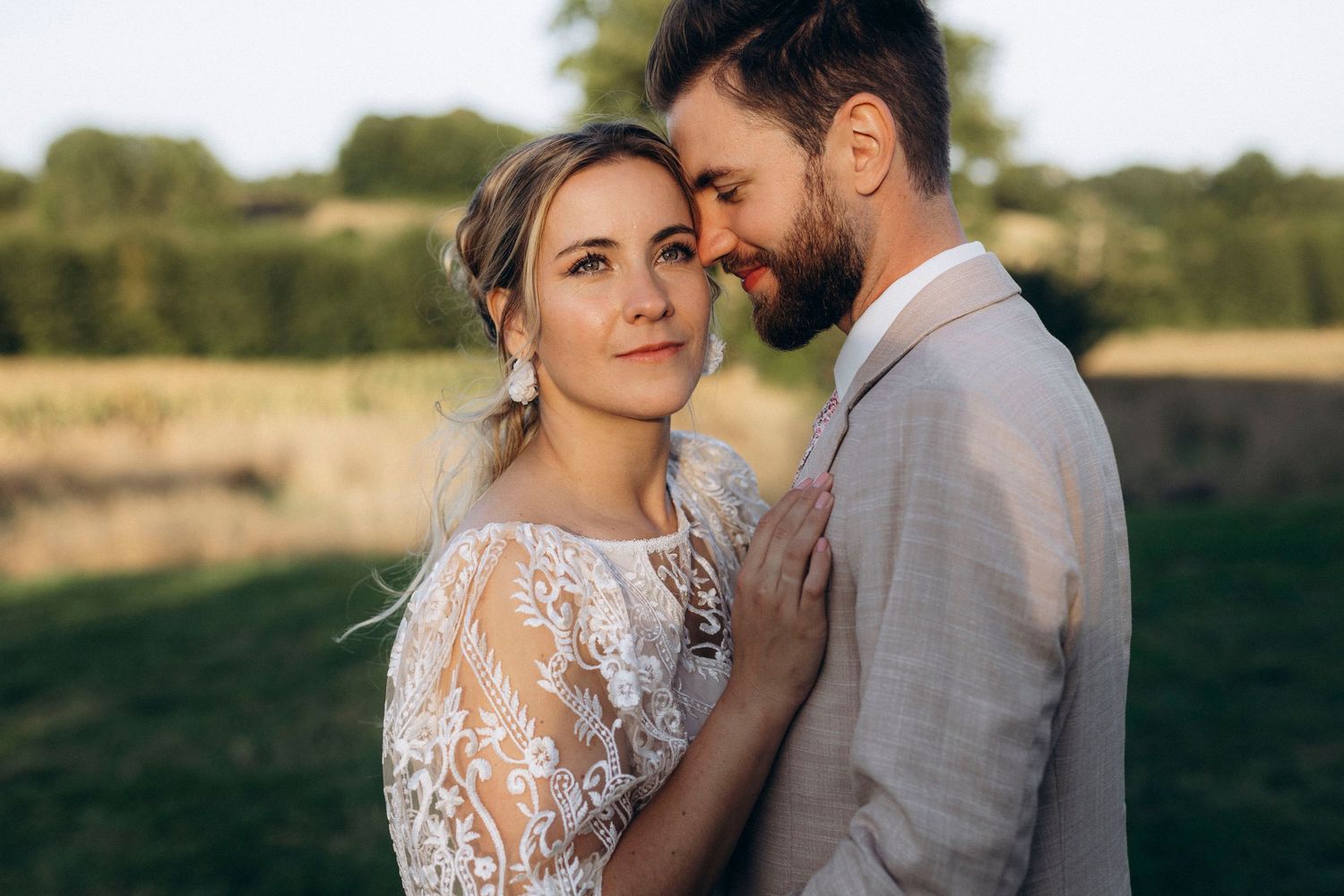 A romantic couple embraces in an outdoor field during golden hour wearing elegant wedding attire.