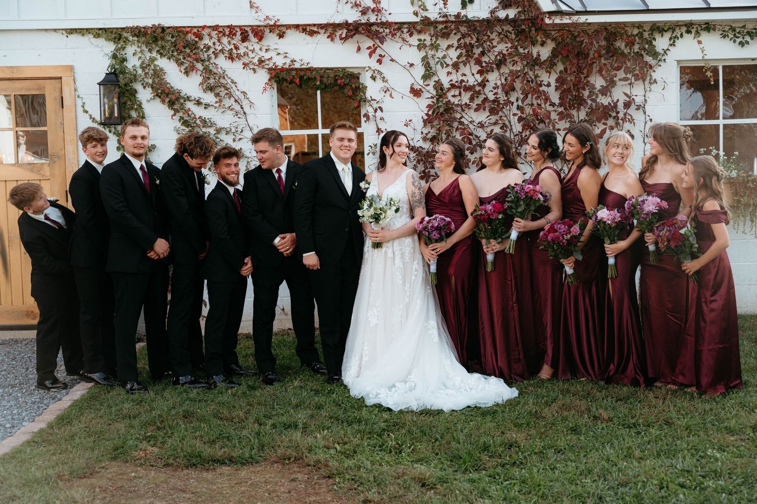 Wedding party members in burgundy dresses and dark suits pose together outdoors on a sunny autumn day.
