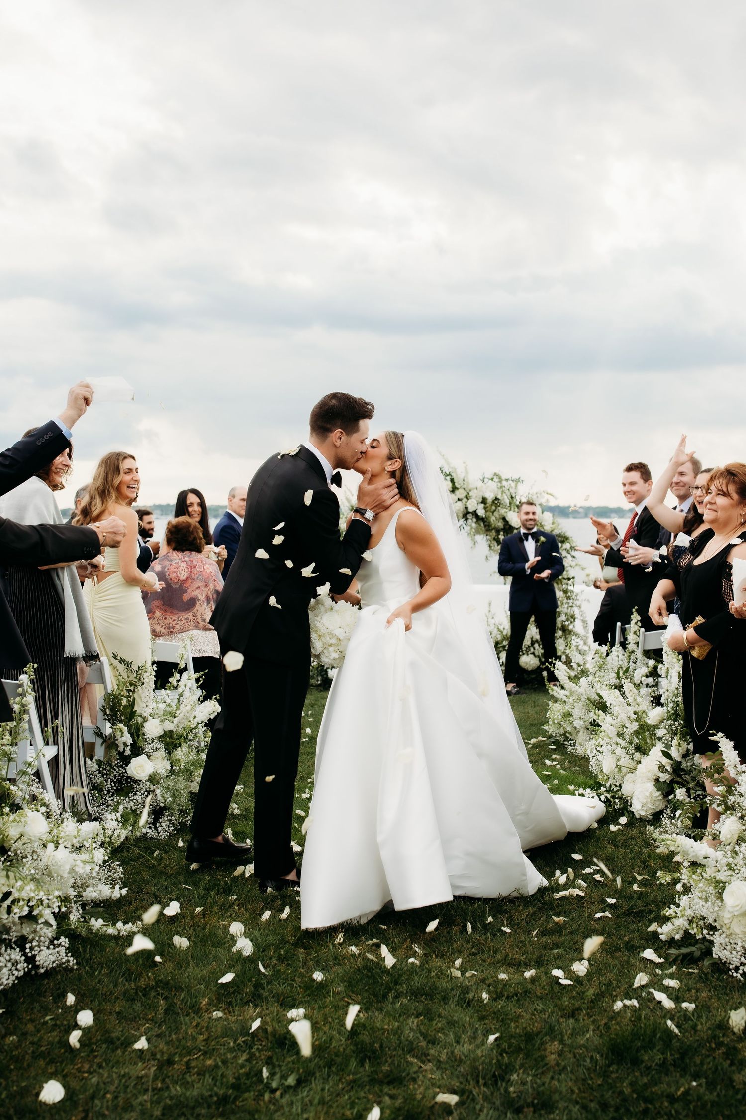 A romantic outdoor wedding ceremony with confetti falling as a couple shares a kiss among guests and floral arrangements.