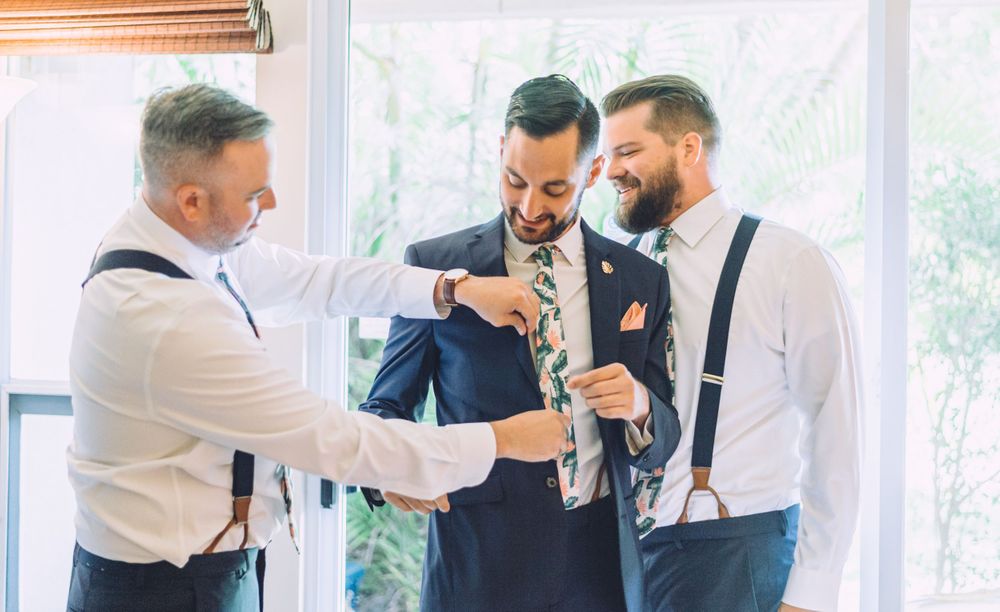 Groomsmen helping with formal attire preparations in bright sunlit room.