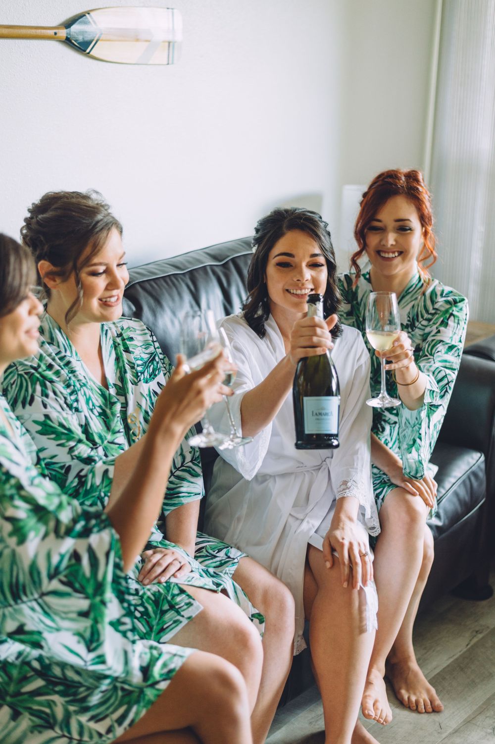 Friends in matching floral robes celebrating with champagne on a grey couch.
