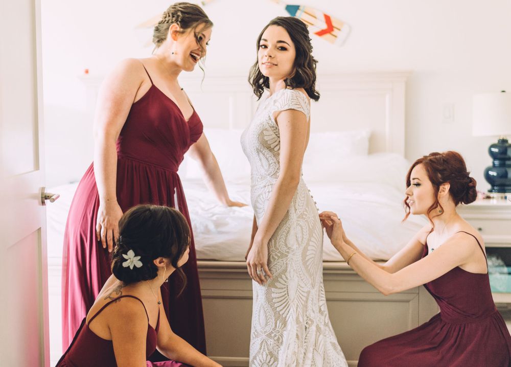 Wedding preparation scene with bridesmaids in burgundy dresses helping with a white gown.