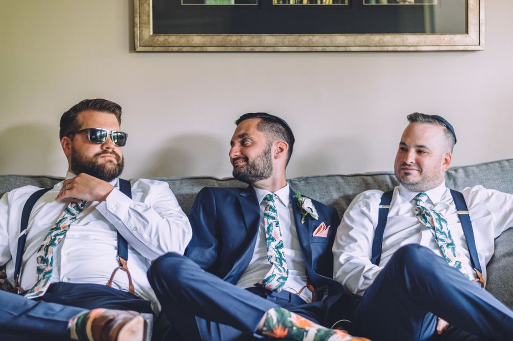 Three groomsmen in matching floral ties relax on a couch during wedding preparations.