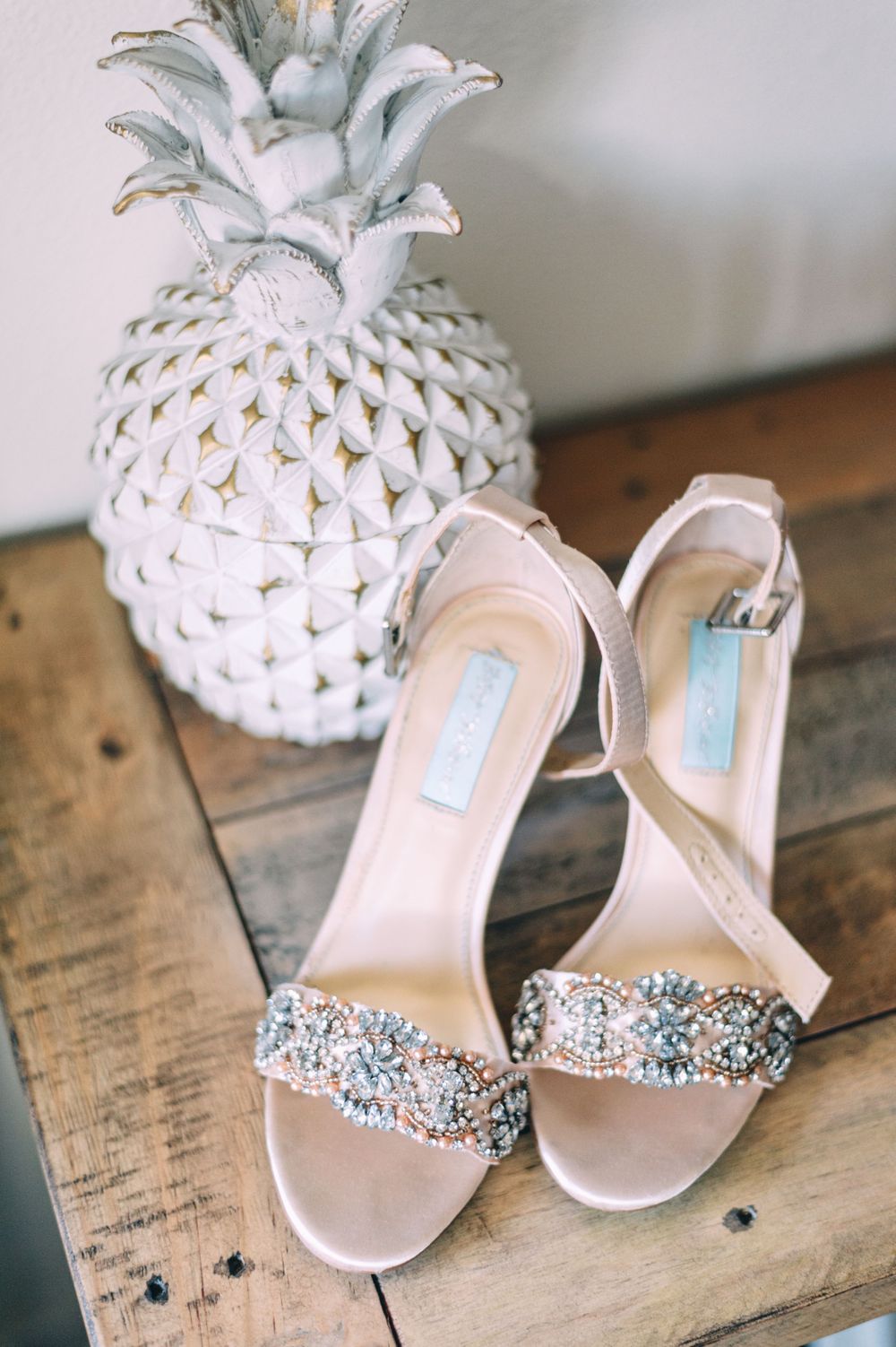 Decorative white ceramic pineapple next to crystal-embellished bridal sandals on wooden surface.
