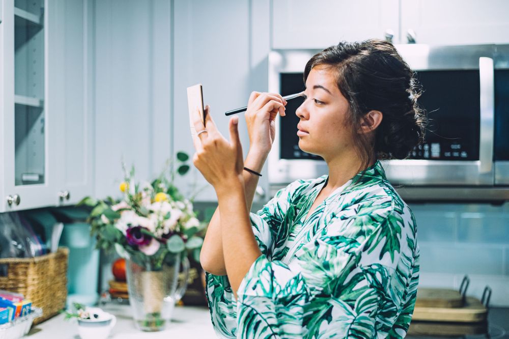 Person applying makeup in a tropical print robe while getting ready in a bright kitchen setting.