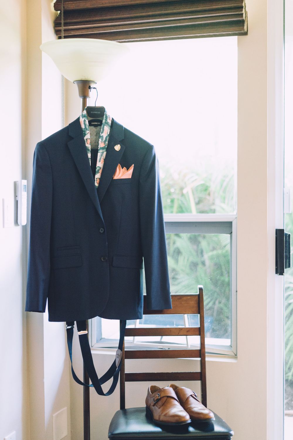 Navy suit jacket with coral pocket square hanging by window with brown leather shoes below.