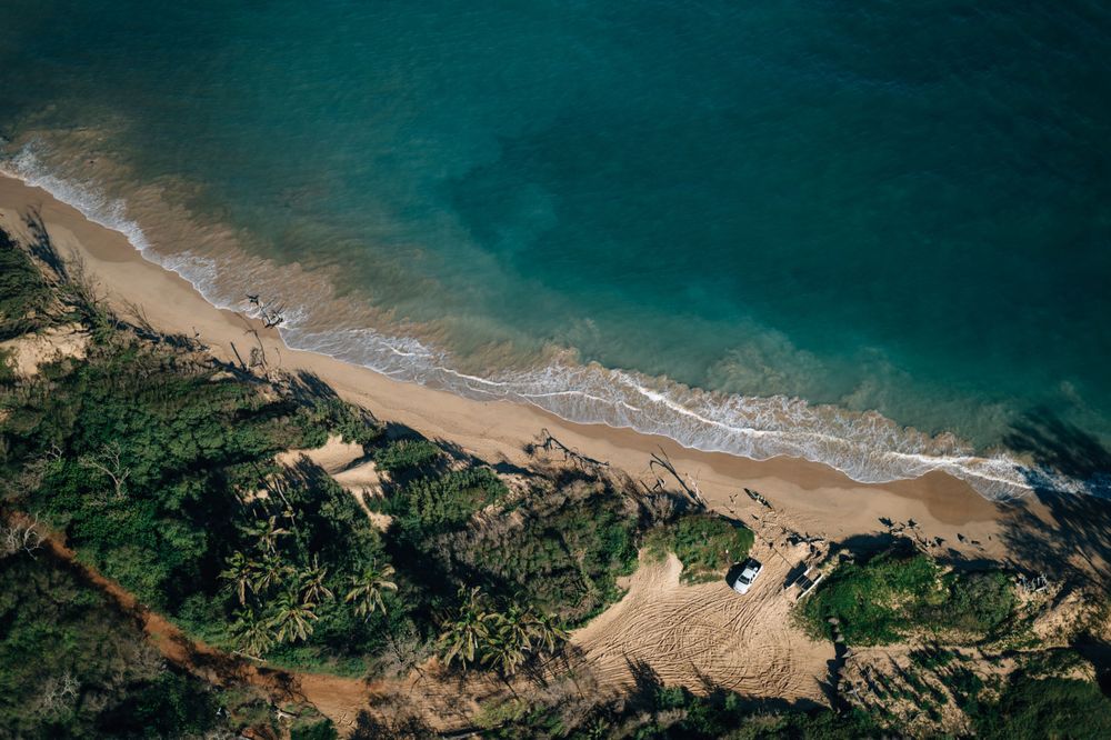 Aerial view of sandy beach and coastline with turquoise waters and vegetation.