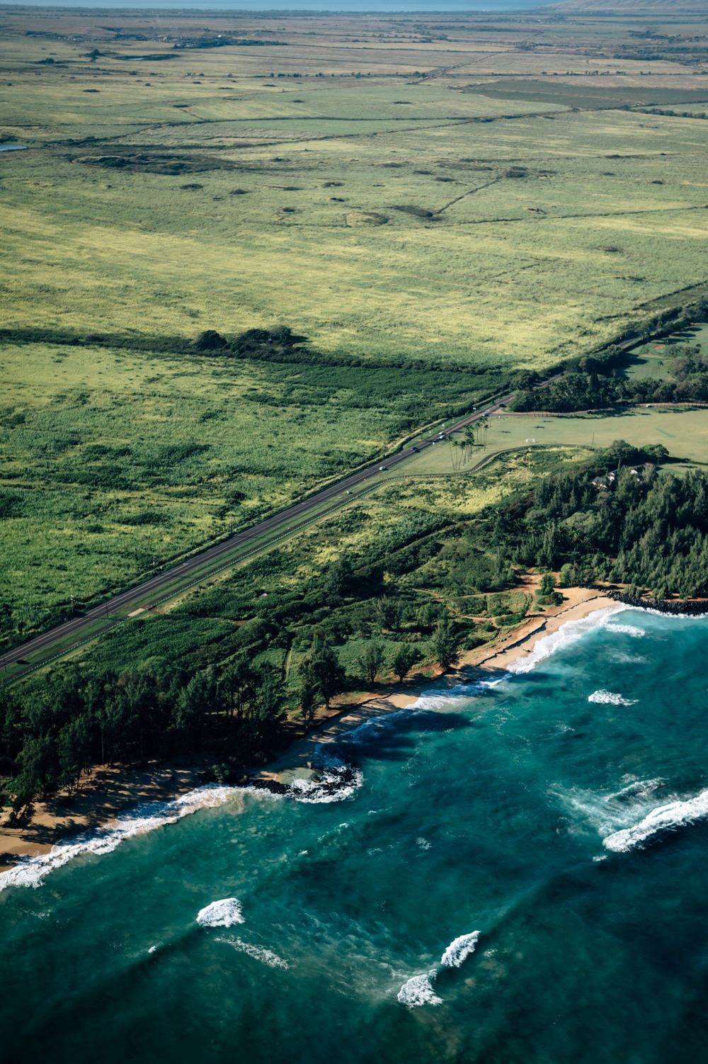 Aerial shot of coastal highway cutting through green fields toward rocky beach and turquoise waters.