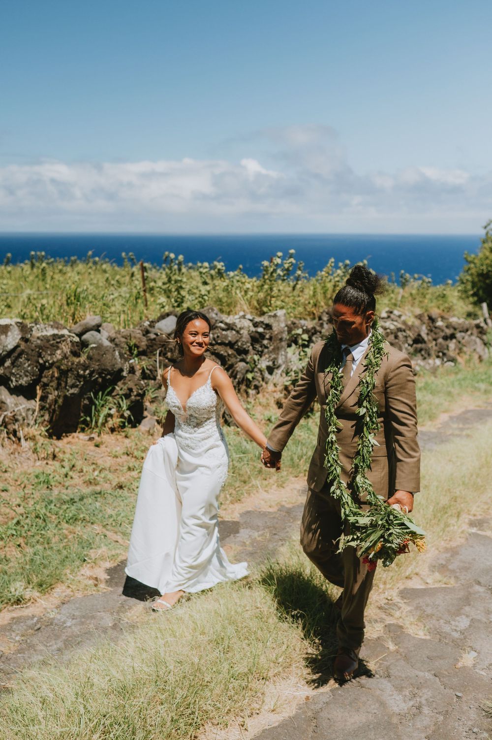 Wedding couple walking along coastal path with ocean views and tropical vegetation.