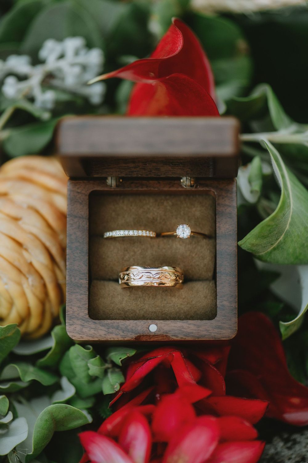 Wedding rings displayed in wooden box surrounded by red roses and green foliage.