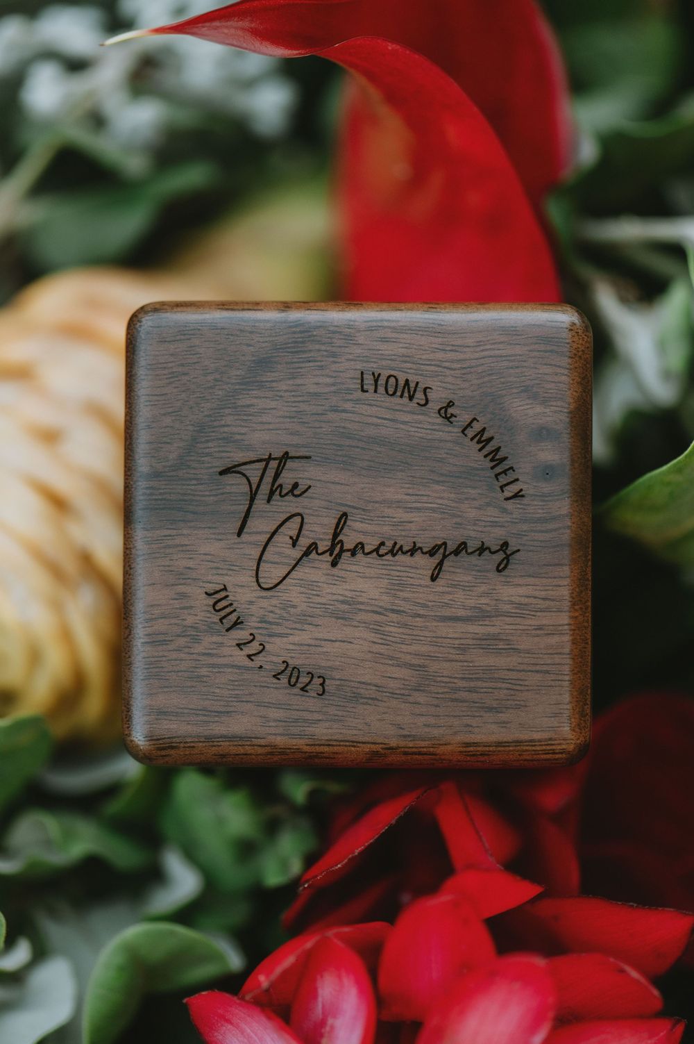 Wooden wedding ring box with engraved names and date surrounded by red and cream colored flowers.
