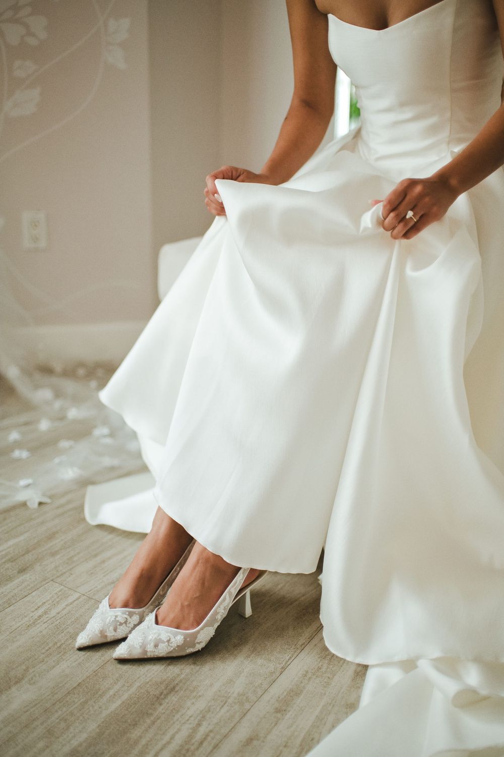 A bride in a white wedding gown adjusts her dress while wearing elegant lace kitten heel shoes on a wooden floor.