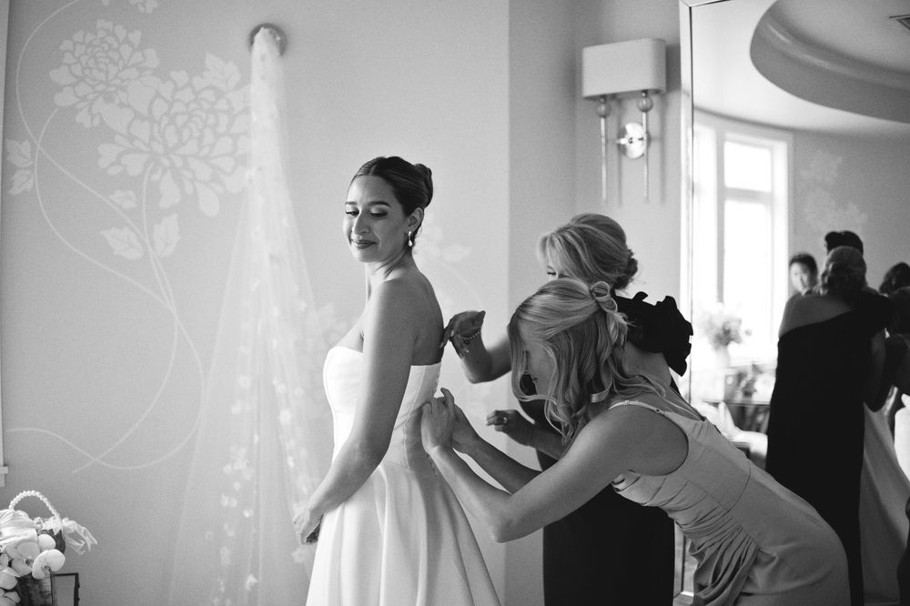 A black and white photograph captures a bride getting ready in a sunlit room as helpers assist with final preparations.
