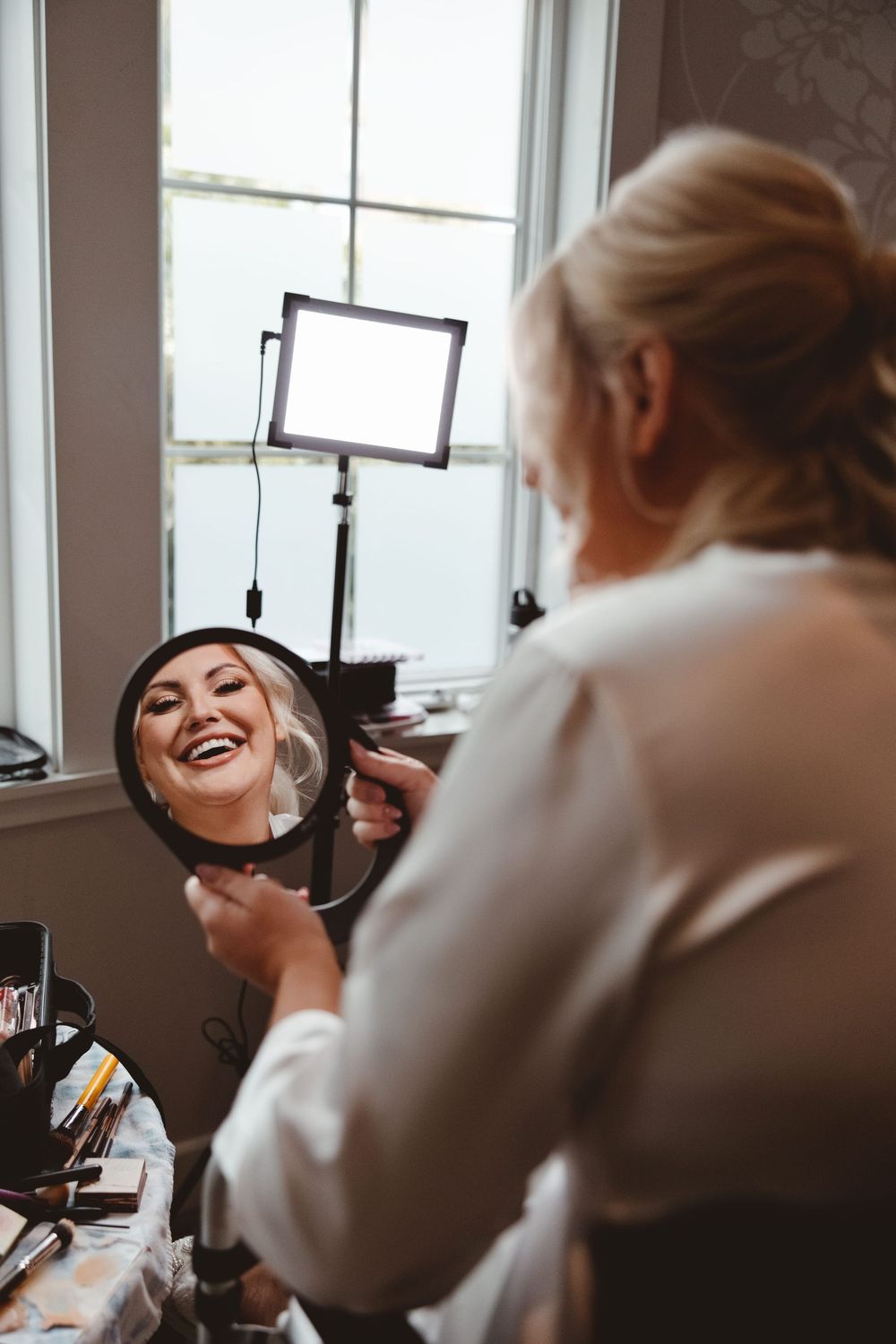 A makeup artist applies cosmetics in a bright, naturally lit room with white walls and windows.