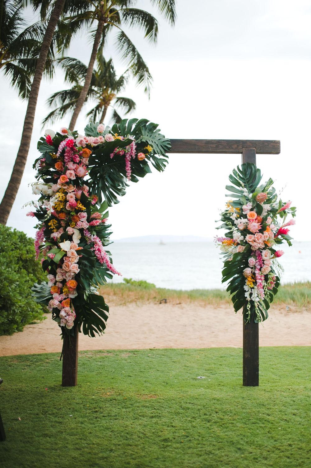A wooden cross decorated with tropical flowers and monstera leaves stands on a grassy beachfront under palm trees.