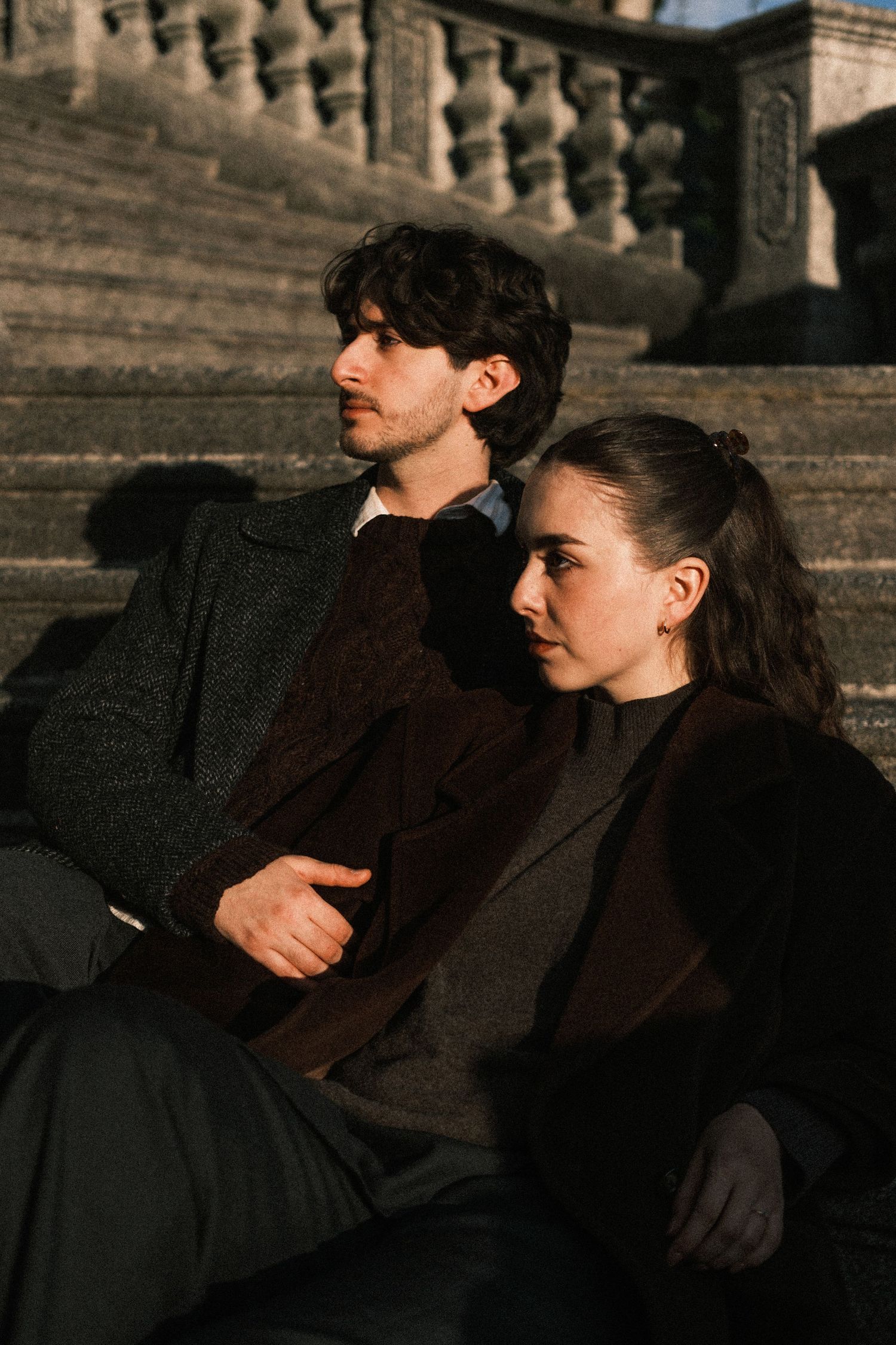 Two figures sit together on stone steps at dusk, silhouetted against an architectural backdrop in warm lighting.