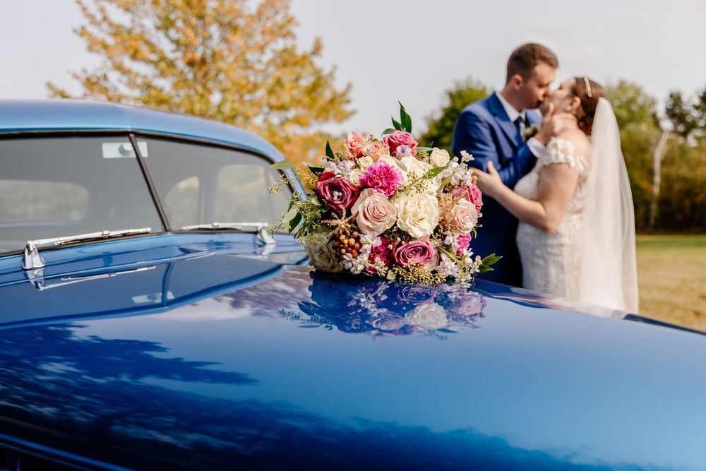 Wedding bouquet of pink and cream roses rests on vintage blue car hood as bride and groom kiss behind it.