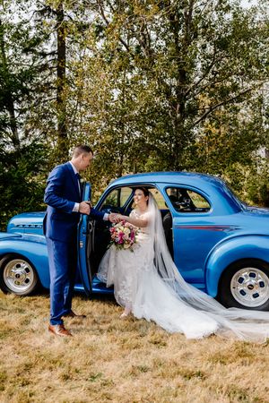 Groom in blue suit helps bride in lace gown exit a classic vintage blue car outdoors.