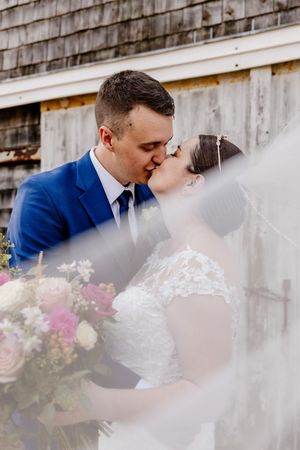 Bride and groom share a romantic kiss framed by flowing veil near a rustic wooden barn.