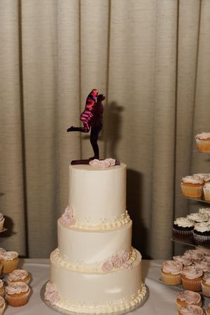 Elegant three-tier white wedding cake with floral details topped with a dancing couple figurine beside cupcakes.