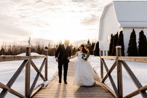 Bride and groom walking hand-in-hand on snowy wooden bridge at sunset near white barn wedding venue.
