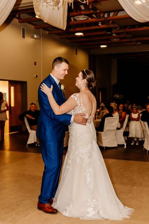 Bride and groom share their first dance in a warmly lit reception hall with guests watching.