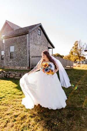 Bride in flowing white gown twirling with sunflower bouquet outside rustic wooden building.