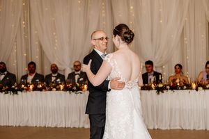 Bride and father share an emotional first dance at elegant candlelit wedding reception with draped white fabric backdrop.