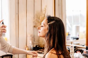 Hairstylist spraying setting spray on bride with long dark hair during wedding preparations.