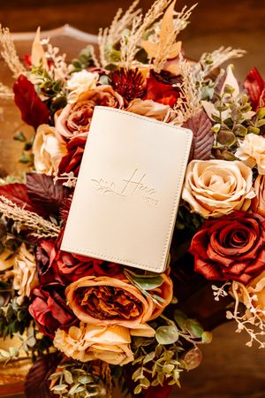 White leather Bible resting on a bouquet of red, burgundy, and champagne roses with greenery.