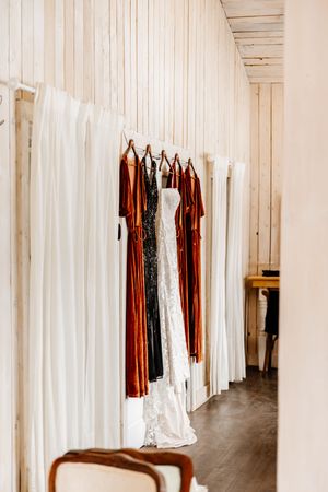 White wedding gown and rust-colored bridesmaid dresses hanging on a rustic white wood wall.