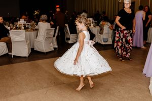 Young flower girl twirls in white dress on wedding reception dance floor, spinning barefoot with joy.