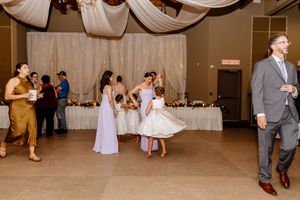 Bridesmaids in lavender dresses dance with flower girl at elegant wedding reception with draped ceiling decor.