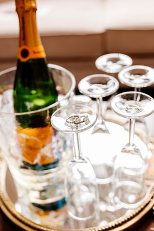 Wedding rings resting on champagne flutes beside a chilled bottle on a gold mirrored tray.