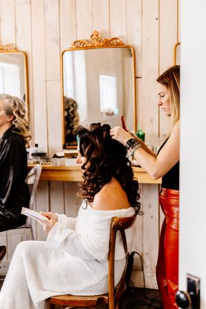 Hairstylist styling bride's dark curly updo in a rustic salon with gold-framed mirror.