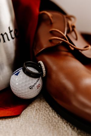 Wedding rings resting on TaylorMade golf ball beside brown leather dress shoes.
