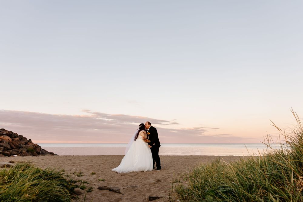 Bride and groom sharing romantic kiss on sandy beach at pastel sunset with ocean backdrop.