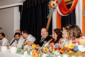 Wedding party seated at decorated head table during colorful fall-themed reception celebration.