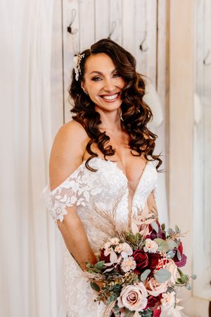 Smiling bride in off-shoulder lace gown holds a stunning red and blush bouquet indoors.