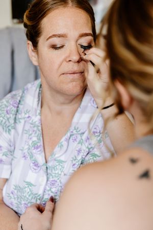 Makeup artist applies eyeliner to a bride wearing a floral robe during wedding day preparations.
