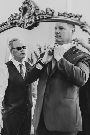 Groomsman helping groom adjust bow tie in front of ornate mirror, black and white.