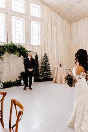 Groom watches bride walk toward him in a light wood ceremony space decorated with greenery.