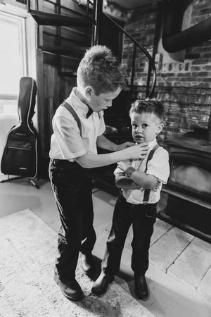 Older boy helps younger brother adjust his suspenders in a black and white wedding preparation photo.