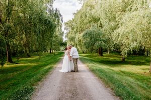 Bride and groom share romantic kiss on gravel path surrounded by lush weeping willow trees.