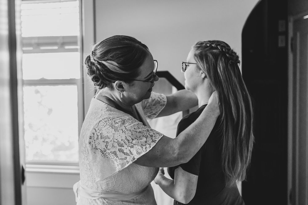 Black and white photo of a woman helping a bride with her dress near a bright window before the wedding.