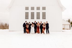 Wedding party posing in front of white barn venue in snowy winter landscape.