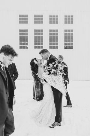Bride and groom share romantic kiss while holding bouquet outside white venue, black and white.