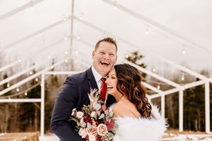 Laughing bride and groom pose together outside a glass greenhouse wedding venue in winter.