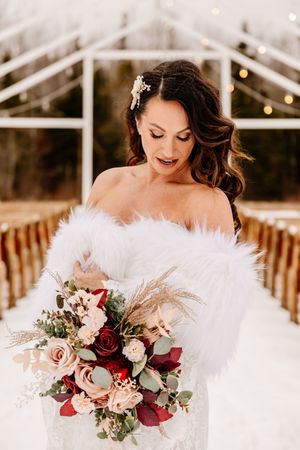 Elegant bride in white lace gown with feather wrap admires her bouquet of red and blush roses.