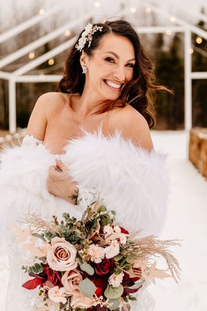 Radiant bride in feather wrap smiles joyfully holding a stunning red and blush floral bouquet.